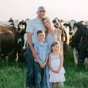 Parents and a little boy and little girl smile while standing in front of a herd of dairy cows